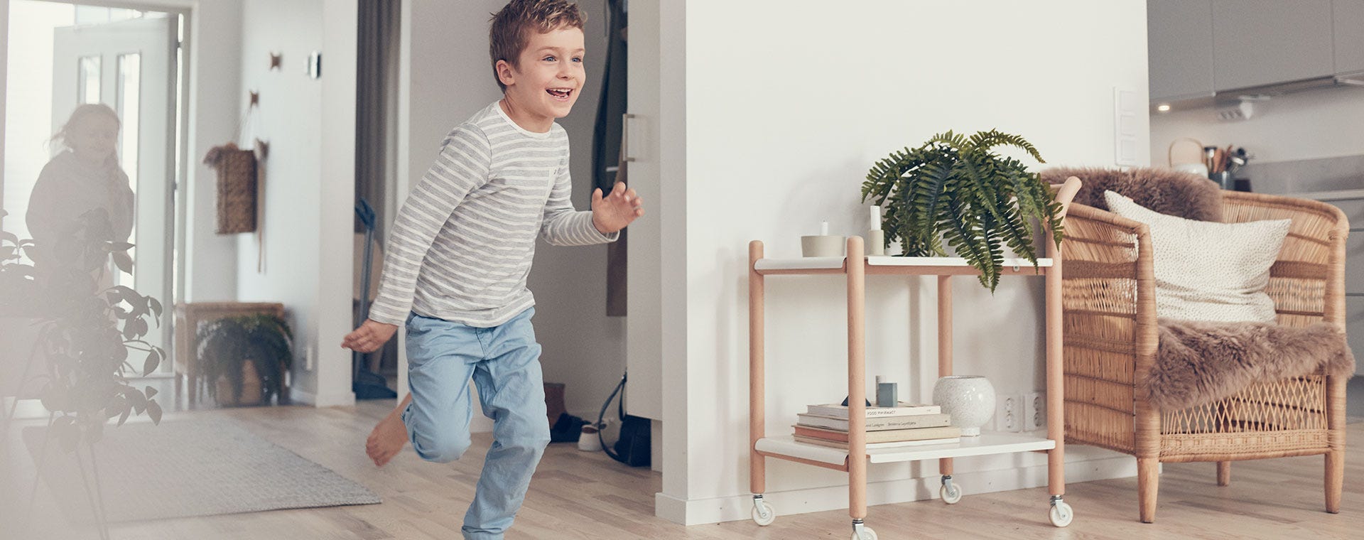 Poika juoksee iloisesti sisällä modernissa ja valoisassa kodissa. Hänellä on yllään raidallinen pitkähihainen paita ja siniset housut. Taustalla näkyy toinen lapsi ovella, sekä vaaleasävyinen sisustus, johon kuuluu rottinkituoli, viherkasveja ja kirjoja. Huoneessa vallitsee lämmin ja kodikas tunnelma.