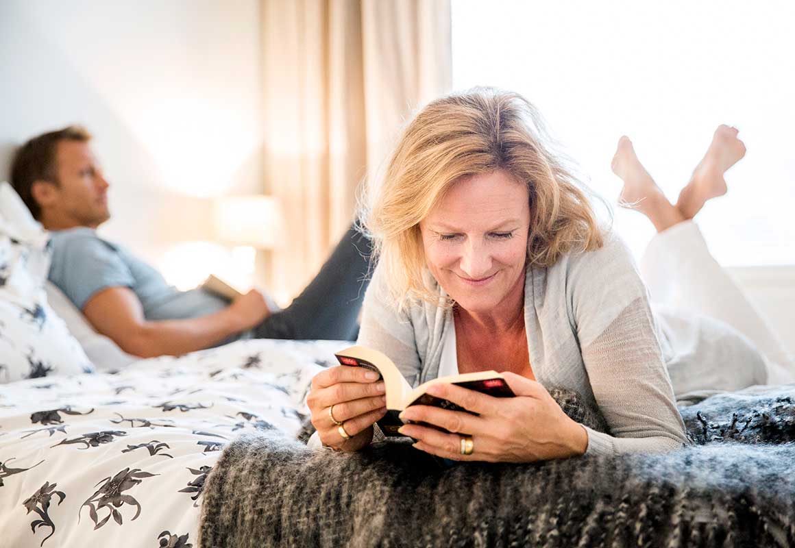 Nainen lukee kirjaa sängyllä rauhallisessa makuuhuoneessa, taustalla mies lepää kodikkaassa ympäristössä.